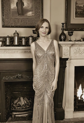 in a 1920s evening dress, standing beside a grand fireplace, with a sepia filter