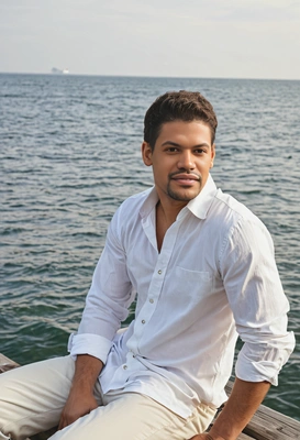 as a man in a loose white shirt and rolled-up pants, sitting on a dock by the sea, looking thoughtfully into the horizon