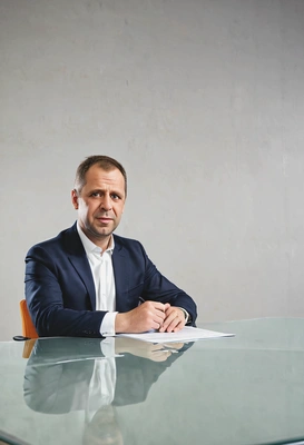 as a CEO, signing a paper, transparent table, sitting on a chair, looking into the camera, blurred background