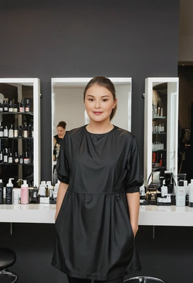 as a hair stylist, wearing a trendy black smock over a stylish outfit, standing in a sleek, modern salon with mirrors and hair products neatly arranged on the counter